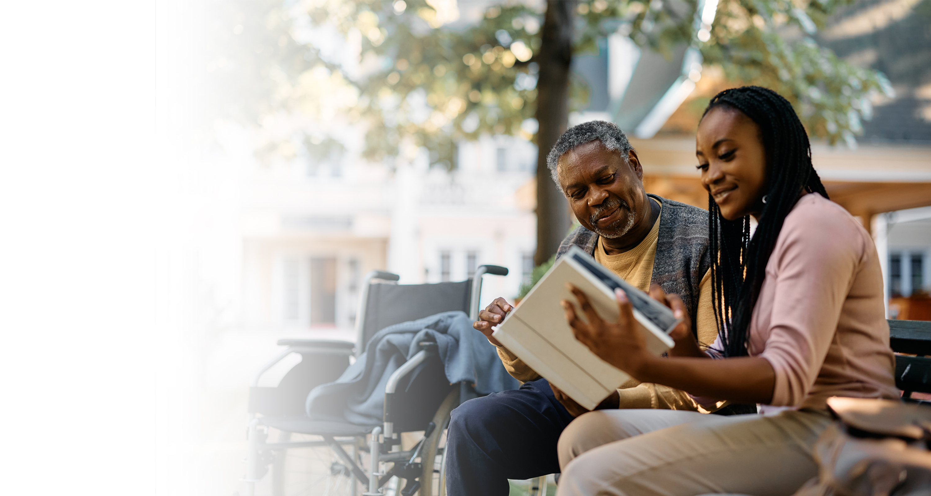 An older adult black man and a younger adult black woman sit on a bench, both smiling and flipping through a photo album together.